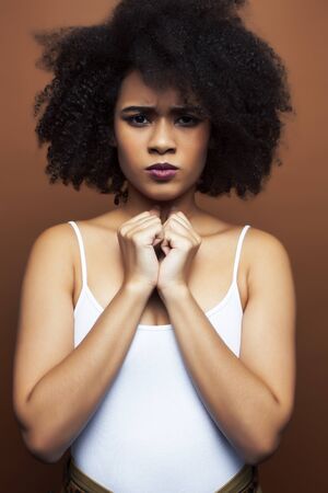 pretty young african american woman with curly hair posing cheerful gesturing on brown background,...