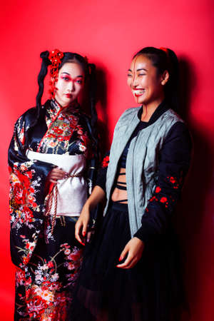 two pretty geisha girls friends: modern asian woman and traditional wearing kimono posing cheerful on red backgroundの写真素材