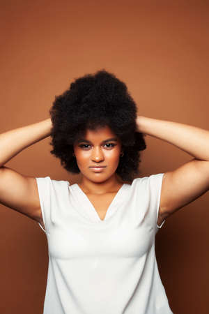 pretty young african american woman with curly hair posing cheerful gesturing on brown background,...