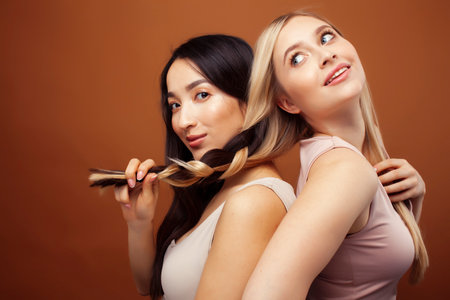 two pretty diverse girls happy posing together: blond and brunette on brown background, asian and caucasian, lifestyle people conceptの写真素材