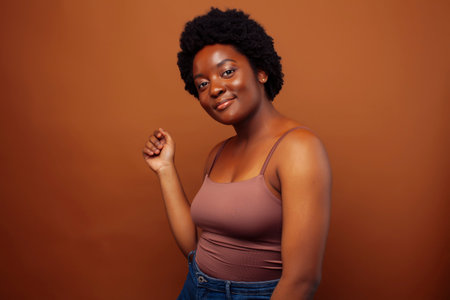 pretty young african american woman with curly hair posing cheerful gesturing on brown background,...
