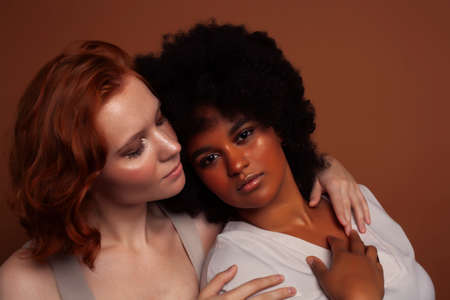 young pretty caucasian, afro woman posing cheerful together on brown background, lifestyle diverse nationality people conceptの写真素材
