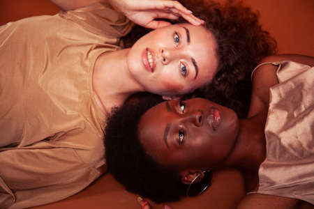 two pretty girls african and caucasian blond posing cheerful together on brown background, ethnicity diverse lifestyle people conceptの写真素材