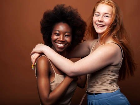 young pretty caucasian, afro woman posing cheerful together on brown background, lifestyle diverse nationality people conceptの写真素材