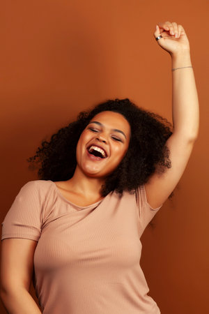 pretty young african american woman with curly hair posing cheerful gesturing on brown background, lifestyle people conceptの写真素材