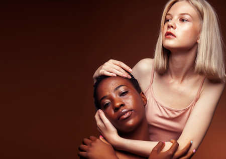 two pretty girls african and caucasian blond posing cheerful together on brown background, ethnicity diverse lifestyle people conceptの写真素材