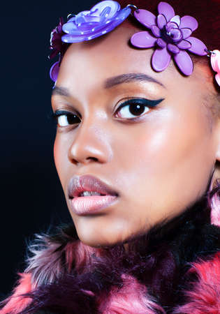 young pretty african american woman in spotted fur coat and flowers on head smiling on black background, fashion people conceptの写真素材