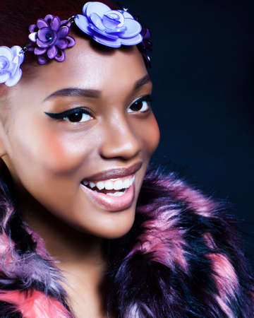young pretty african american woman in spotted fur coat and flowers on head smiling on black background, fashion people conceptの写真素材