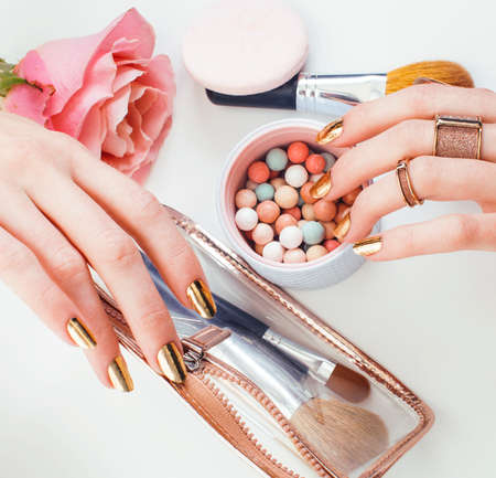 woman hands with golden manicure and many rings holding brushes, cosmetic and rose flower on white background, spa treatment conceptの写真素材
