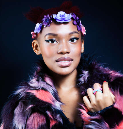 young pretty african american woman in spotted fur coat and flowers on head smiling on black background, fashion people conceptの写真素材