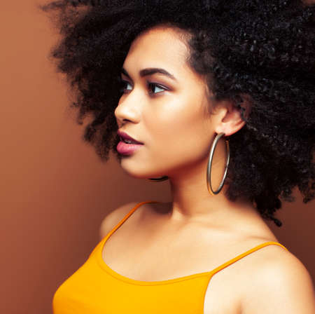 pretty young african american woman with curly hair posing cheerful gesturing on brown background,...