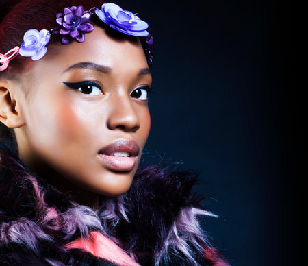 young pretty african american woman in spotted fur coat and flowers on head smiling on black background, fashion people conceptの写真素材