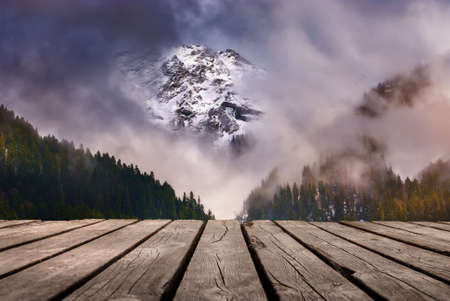 White capped mountain piercing through the clouds and empty wooden table in nature outdoor. Natural template landscape.の写真素材