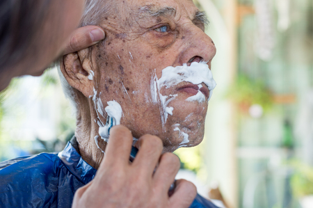 Elderly man getting his beard shaved by young skilled man at homeの写真素材