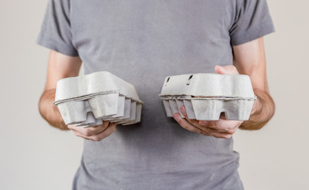 Caucasian man with gray t-shirt holding two cardboard egg boxes full of hen eggs on a white...