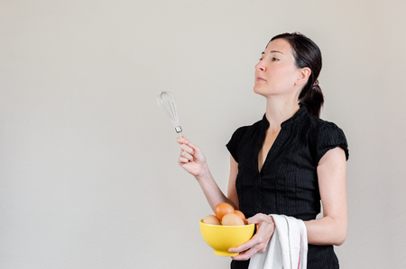 Beautiful Caucasian woman with black shirt holding an eggbeater and a yellow bowl full of hen eggs.の写真素材