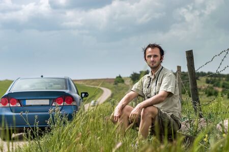 Caucasian man sitting in a rural area near his blue car on a cloudy spring day. Freedom concept.