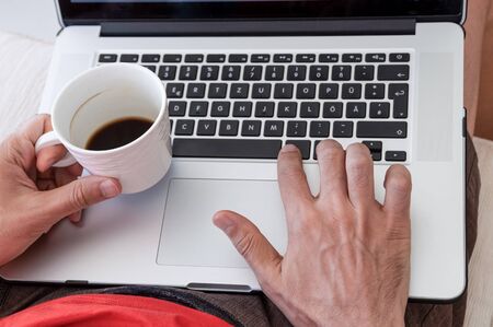 Young businessman holding mug of coffee while working on laptop computer on a couch at home. Portable office concept.の写真素材