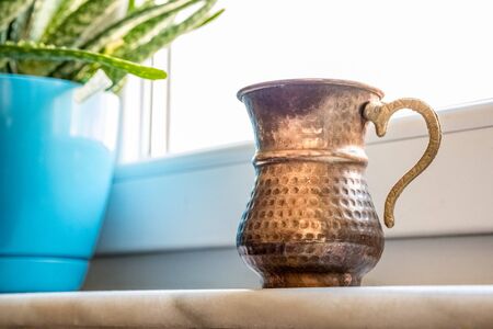Close up of a copper mug used for drinking water, near the aloe vera plant on the window sill.の写真素材