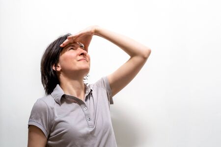 Portrait of a pretty young brunette woman looking far away or watching  with hand over forehead against a white wall.の写真素材