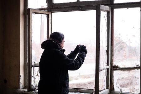 Adult man with black cold winter coat taking pictures of the outside view from the broken window of an abondened house on a cold winter day.の写真素材