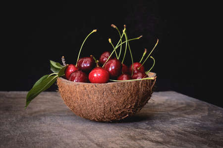Fresh cherries in a coconut shell bowl on a wooden table on black background. Healthy eating.の写真素材