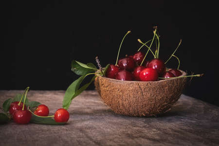 Fresh cherries in a coconut shell bowl on a wooden table on black background. Healthy eating.の写真素材