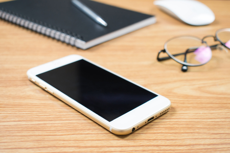 A close up photo of a phone on a brown wooden desk with eyeglasses, notebooks, pens and computer mounts.の写真素材