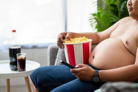 Close-up of an overweight young man sitting on the TV and holding the remote control on the sofa in...