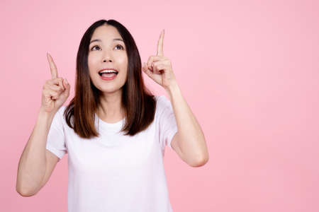 Portrait of a happy excited asian woman standing and pointing two fingers up at copy space isolated over pink background.の写真素材