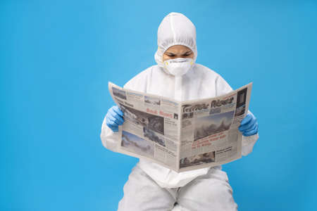 Asian woman in protective clothing with gloves and mask. She is reading a newspaper about COVID-19.の写真素材