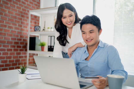 Young Asian couple is using a laptop while shopping at an online website at home. They are happy to enjoy the weekend.の写真素材
