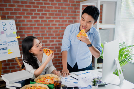 Young Asian colleague is eating pizza and exchanging ideas on his desk. They are happy in the office.の写真素材