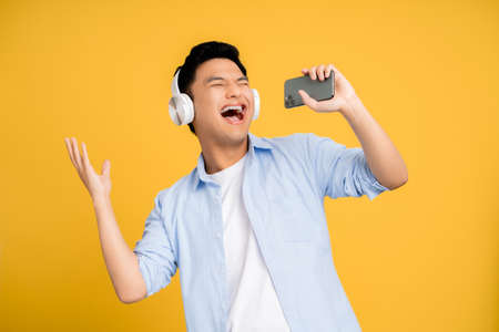 Young Asian man enjoys singing his favorite song. He is wearing wireless headphones. Yellow background.の写真素材