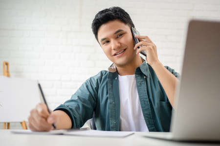 Portrait of a young Asian man in casual clothes sits at a white desk. He is taking notes and talking on the phone.の写真素材