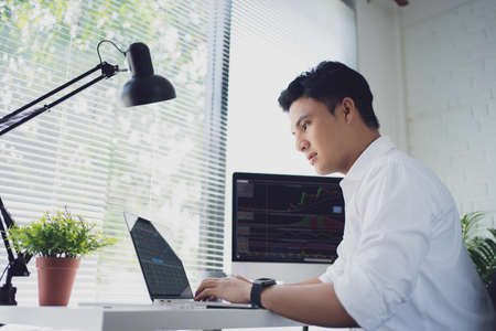 Young Asian man trades stocks on his laptop screen on his desk. He is serious about trading stocks.