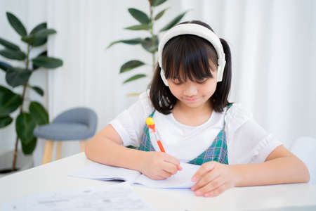 Child wearing headphones and studying online via video call at home in the situation of spreading the corona virus. She is writing a book.の写真素材