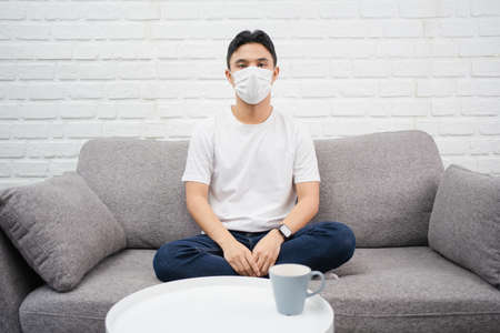 Young asian man staying at home wearing mask sitting on couch. He home isolation.の写真素材
