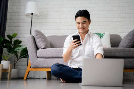 Happy young asian man using mobile phone while sitting at a couch at home with laptop computerの写真素材