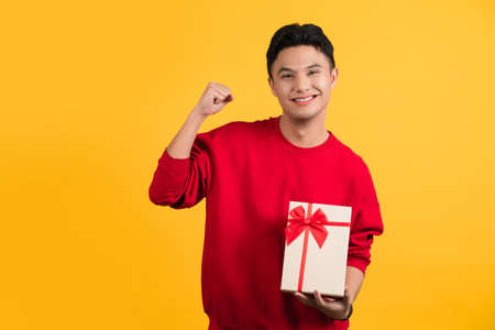 Handsome young man wearing red T-shirt holding gift box isolated on yellow background studio portrait.の写真素材