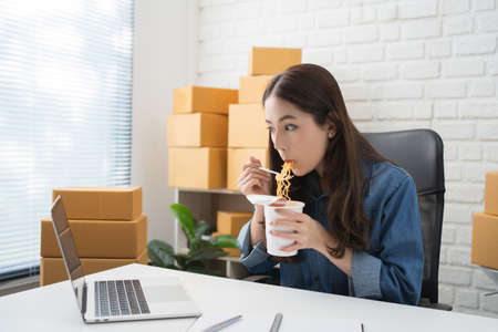 Asian business woman eating instant noodles while working on laptop computer at home office, Happy...
