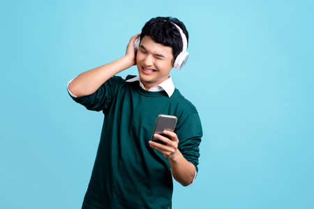 Portrait of smiling young handsome Asian man wearing headphones listening to music from smartphone...