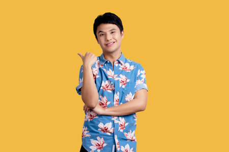 Young Asian man in blue floral shirt smiling and pointing hand sideways on yellow background.の写真素材
