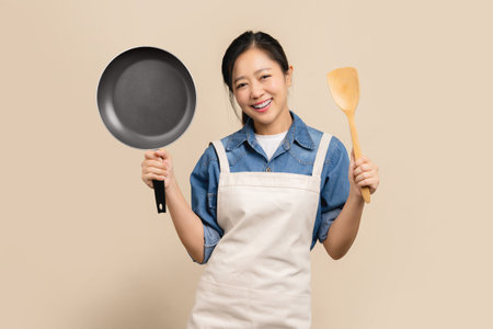 Young Asian housewife in apron and holding pan standing isolated on light brown background.の写真素材