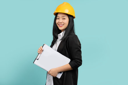 Portrait of happy smiling woman in yellow helmet and black suit She looks at the camera while standing and holding a clipboard with documents isolated over a green background.の写真素材