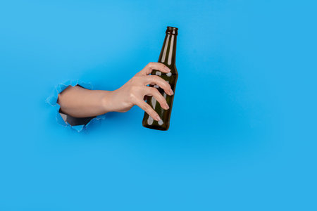Hand through paper holding alcohol bottle on blue backgroundの写真素材
