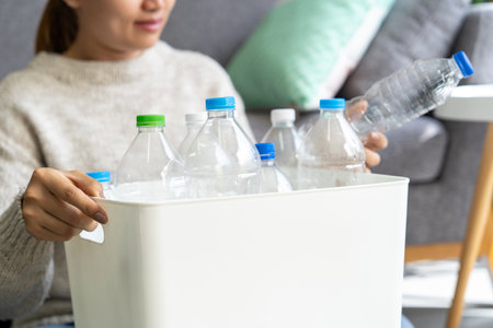 Happy Asian woman wearing a cream-colored sweater collects and sorts recycled plastic bottles into...