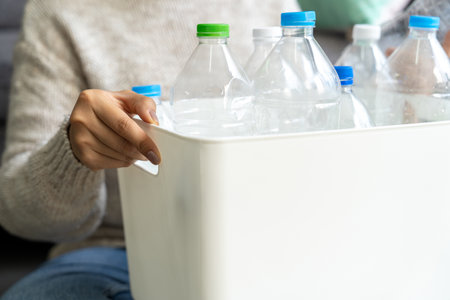 A woman wearing a cream sweater collects and sorts recycled plastic bottles into white trash cans in a living room.の写真素材