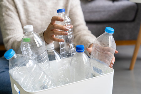 A woman wearing a cream sweater collects and sorts recycled plastic bottles into white trash cans.の写真素材