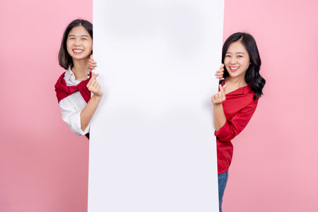 Two beautiful Asian girls standing behind the white blank banner or empty copy space advertisement board and showing mini heart signの写真素材